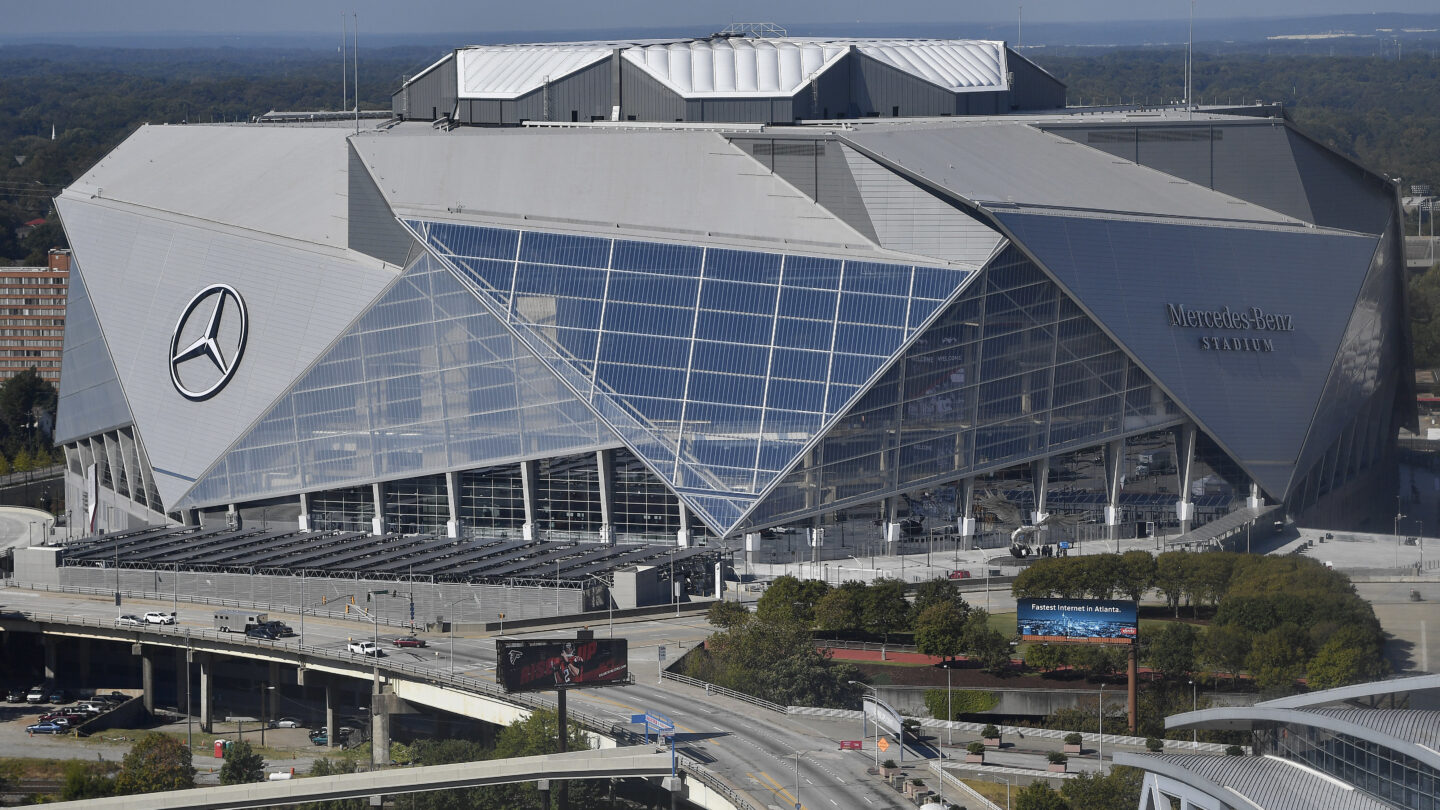 A wide photo of the entire Mercedes-Benz stadium in Atlanta. A giant Mercedes-Benz logo is on the side of the gray, angular, geometric building.