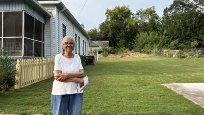 An older lady stands in front of a short, stone wall and green lawn laid with fresh sod. Her house is to the left, a gray one-story with a short wood picket fence and screened-in porch.