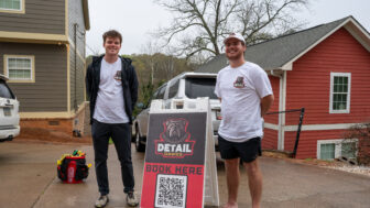 Two guys standing with a sign that says Detail Dawgs (pronounced dogs) in between them.