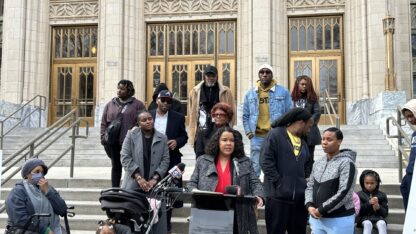 A group of residents from West Atlanta neighborhoods stand on the steps of the Atlanta City Hall. Attorney Meghan Jones in a red shirt and gray overcoat stands at a podium in the center.