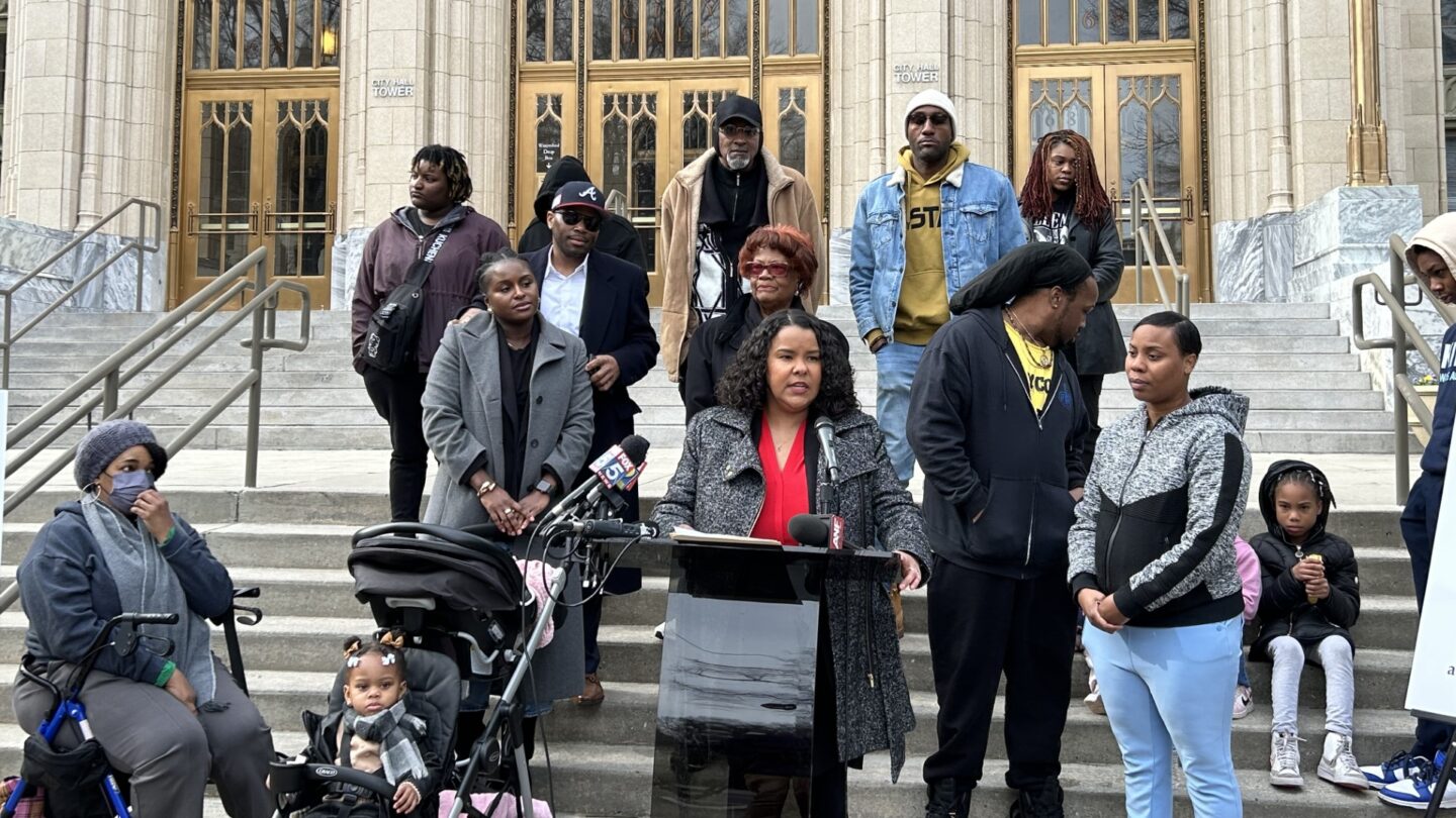 A group of residents from West Atlanta neighborhoods stand on the steps of the Atlanta City Hall. Attorney Meghan Jones in a red shirt and gray overcoat stands at a podium in the center.