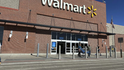 The front of a brown brick Walmart store with the Walmart name and logo, six yellow lines pointing inward in a circle.