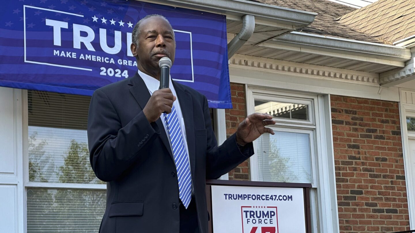 Ben Carson wears a suit and holds a microphone as he speaks at a ceremony to open Donald Trump's first Georgia campaign office on June 13. Behind Carson hangs a blue campaign banner.