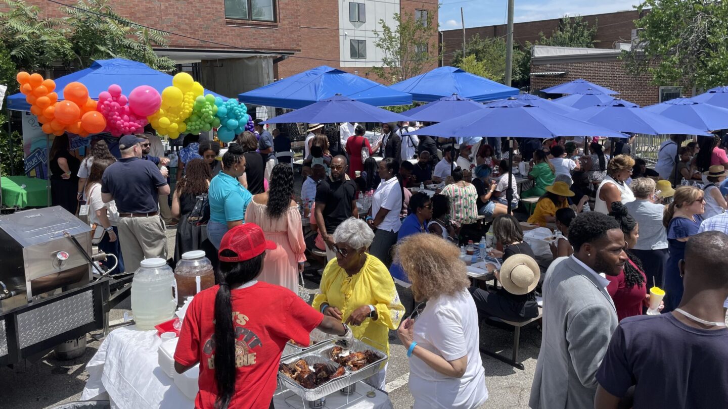 People eat food outdoors under blue patio umbrellas at a Juneteenth block party to mark the opening of the Biden campaign's Atlanta office.