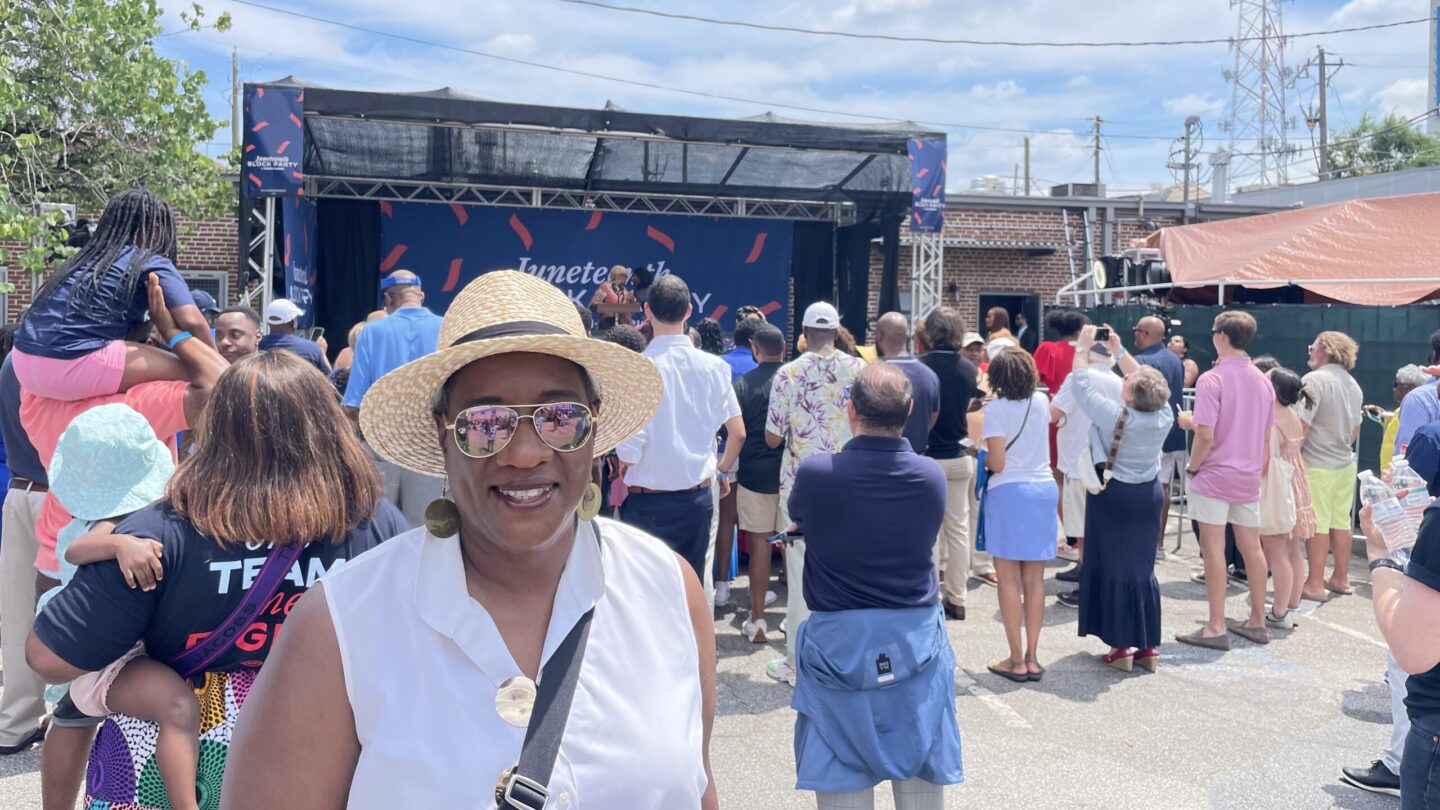 Wearing sunglasses, a straw hat and a sleeveless white shirt, Val Acree attends a Juneteenth block party. Behind her is an outdoor stage, with many people standing in front of the stage, facing it.