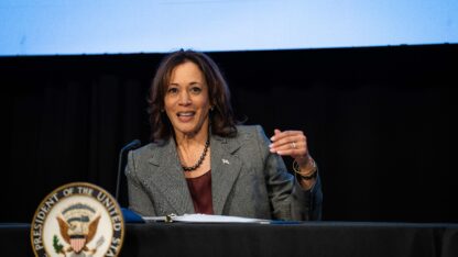 Vice President Kamala Harris delivers remarks while sitting at a table, leaning into a microphone. The seal of the vice president is in front of the table where she sits.