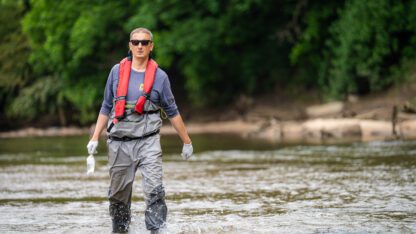 Jason Ulseth, wearing an orange PFD and grey waiters on top of a blue, long sleeve shirt wades through knee height water in the Chattahoochee.