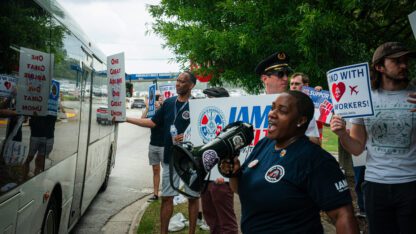 Union members and supporters stand on a sidewalk, holding signs and chanting as a Delta bus passes by them.