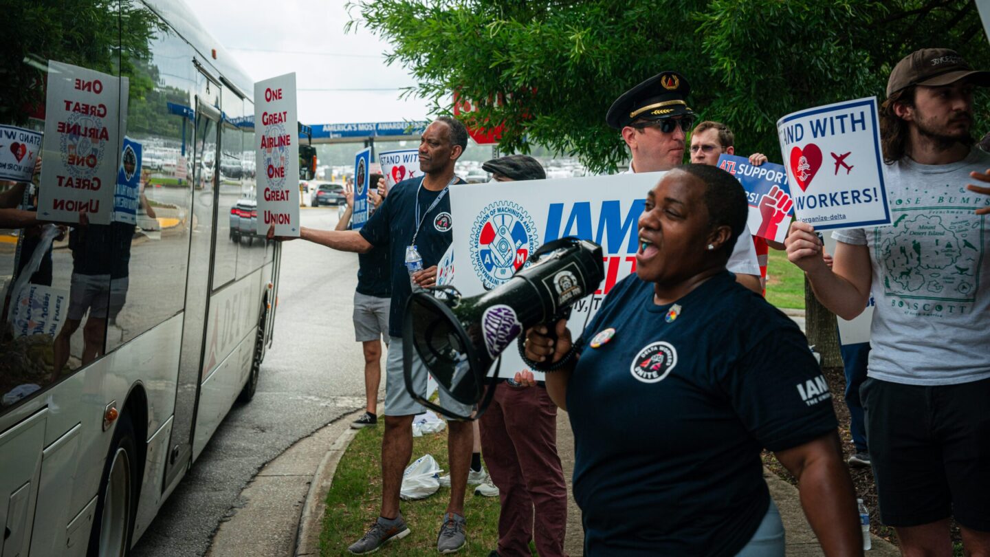 Union members and supporters stand on a sidewalk, holding signs and chanting as a Delta bus passes by them.