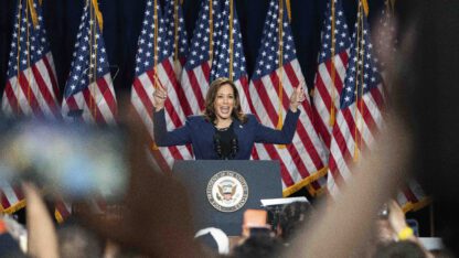 Vice Kamala Harris raises her hands at a rally at West Allis Central High School. A row of American flags stand behind her.