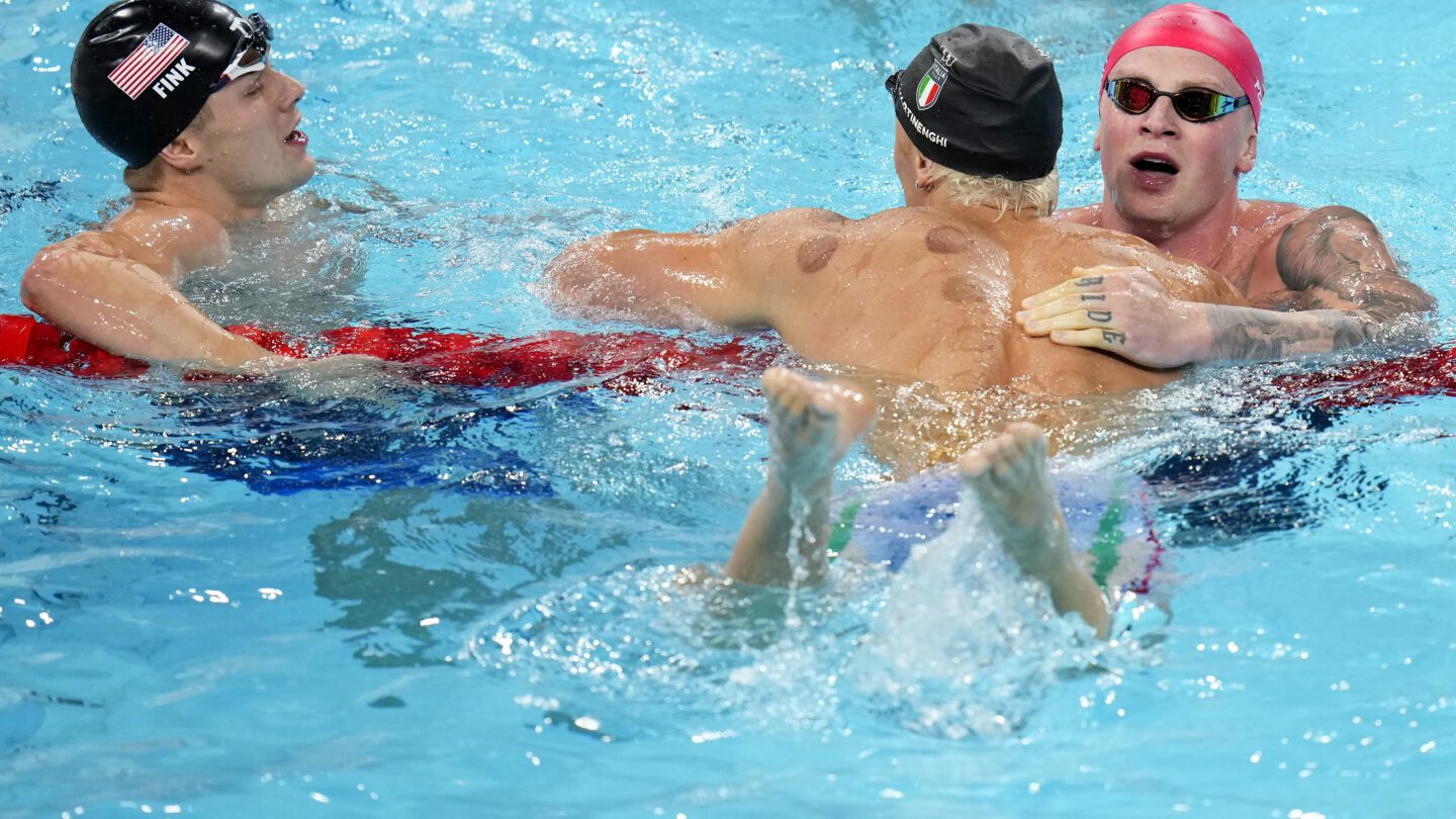 Nicolo Martinenghi, center, of Italy, is congratulated by Adam Peaty, of Great Britain, right, and Nic Fink, of the United States, left, after winning the men's 100-meter breaststroke final at the 2024 Summer Olympics on Sunday. All three men are hovered over the lane line in the pool, cap and goggles still on.