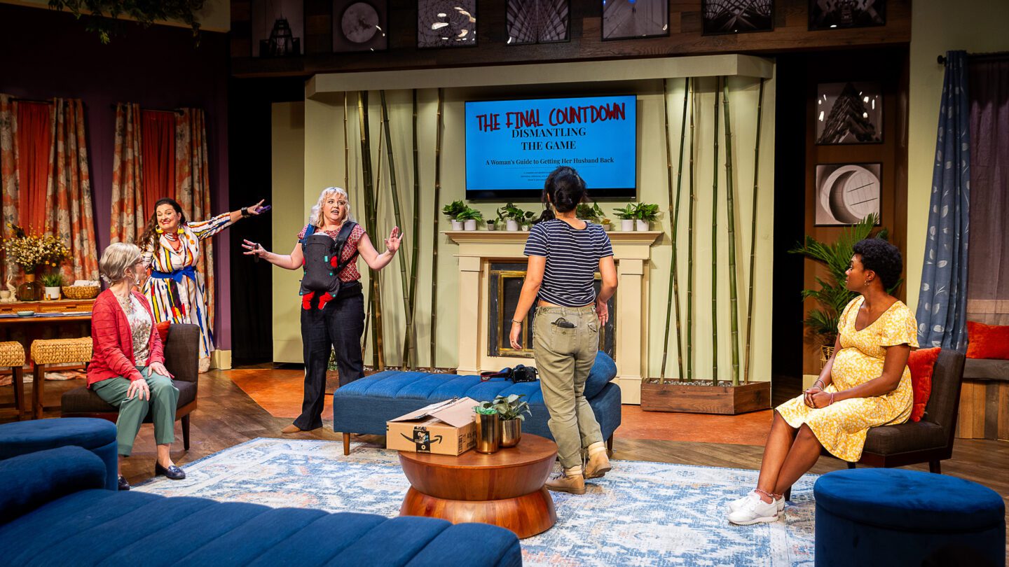 Actors perform on the set of a play in front of a backdrop featuring a fireplace and a TV displaying the words 