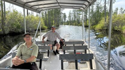 Two men sit on a 24 foot jon boat that has two rows of benches facing the front of the boat. It has a metal cover in top of the boat. In the background is a canal of dark water and green trees on both sides.