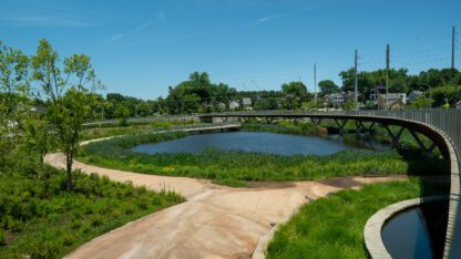 Cook Park. A cement path bordered by grass leads to a blue pond. The sky is blue and cloudless.