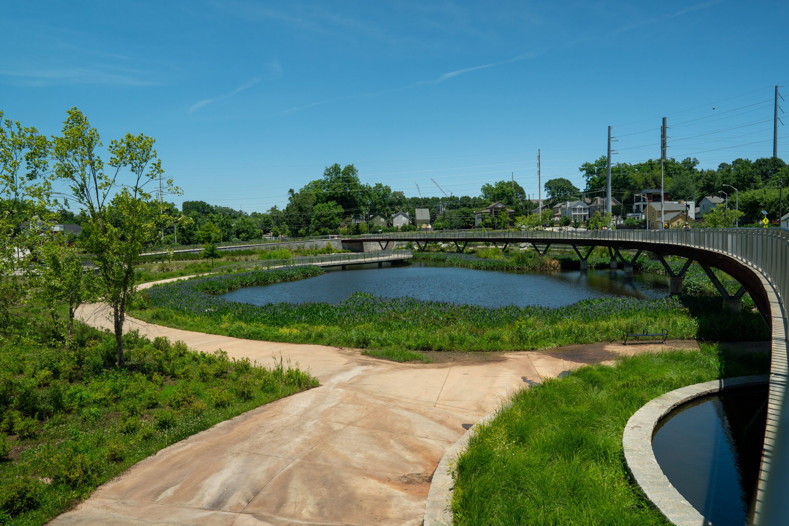 Cook Park. A cement path bordered by grass leads to a blue pond. The sky is blue and cloudless.