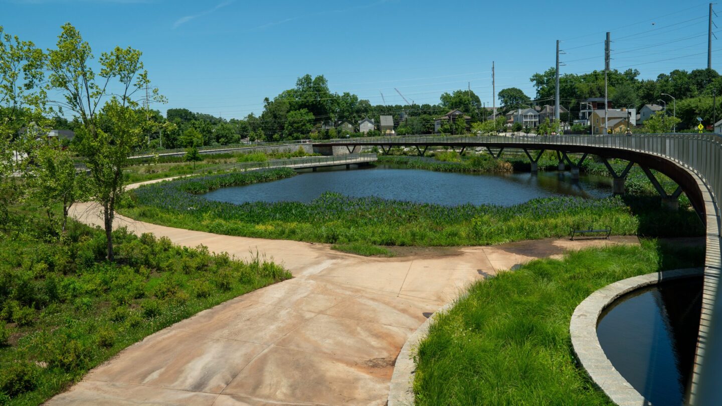 Cook Park. A cement path bordered by grass leads to a blue pond. The sky is blue and cloudless.