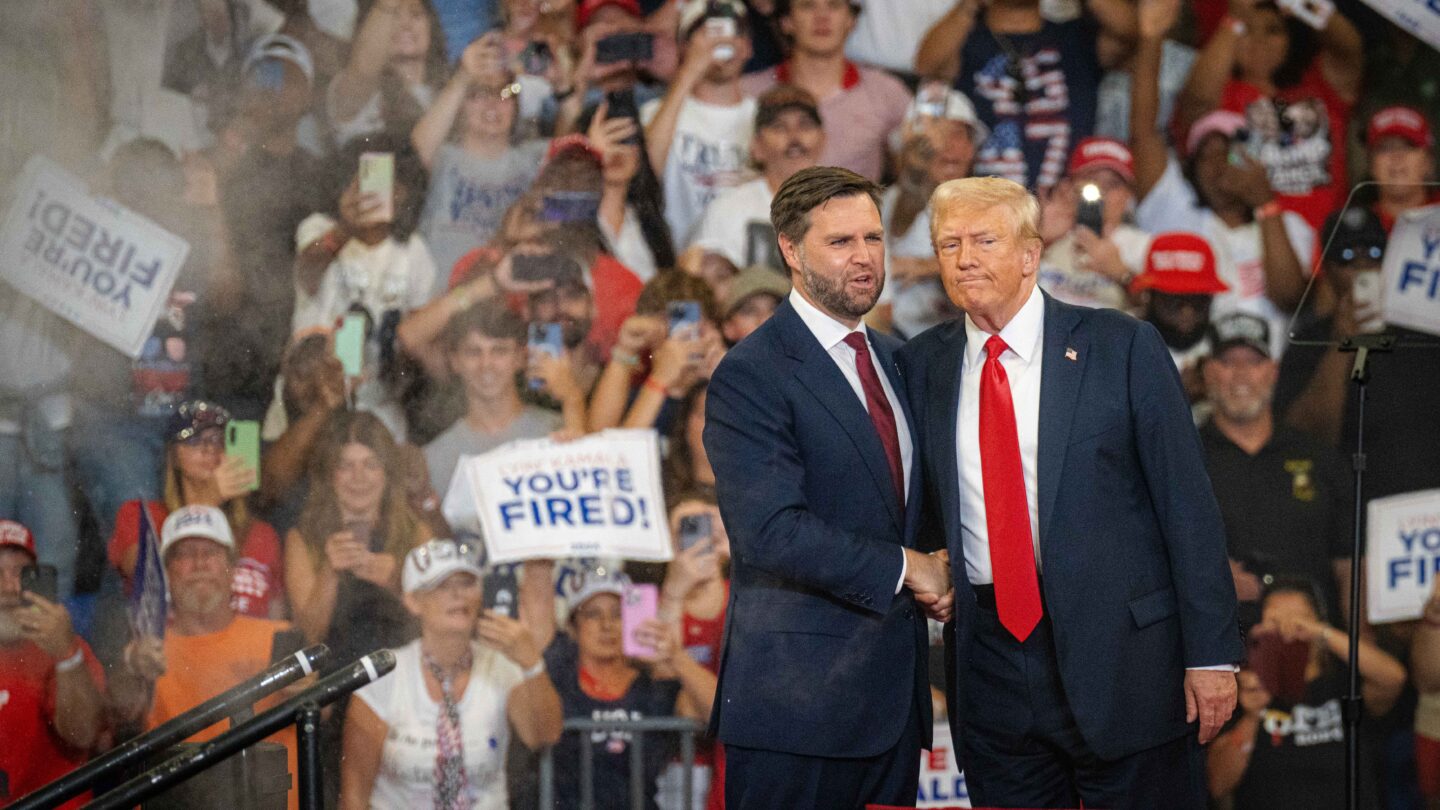 Ohio Sen. JD Vance stands to the left of former President Donald Trump on stage with a crowd behind them.