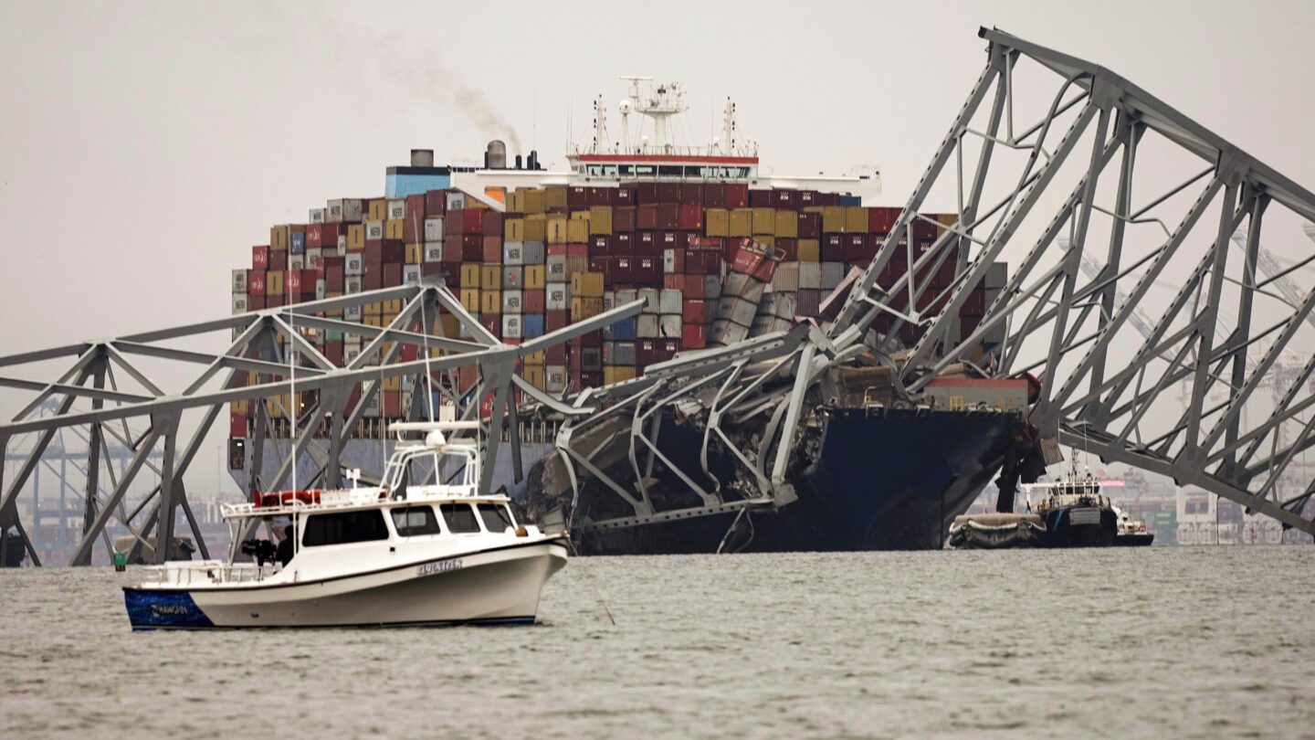 A small boat is anchored in front of a cargo ship that is stuck under the part of the structure of the Francis Scott Key Bridge after the ship hit the bridge Wednesday, March 27, 2024, in Baltimore, Md.