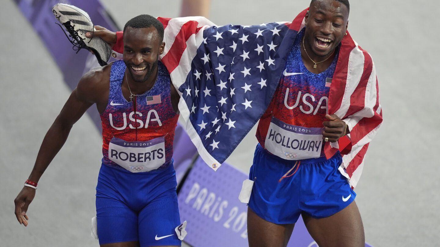 Gold medalist Grant Holloway, of the United States, right, celebrates with silver medalist Daniel Roberts, of the United States, at the end of the men's 110-meter hurdles final, at the 2024 Summer Olympics. Holloway has an American flag draped over his shoulders.