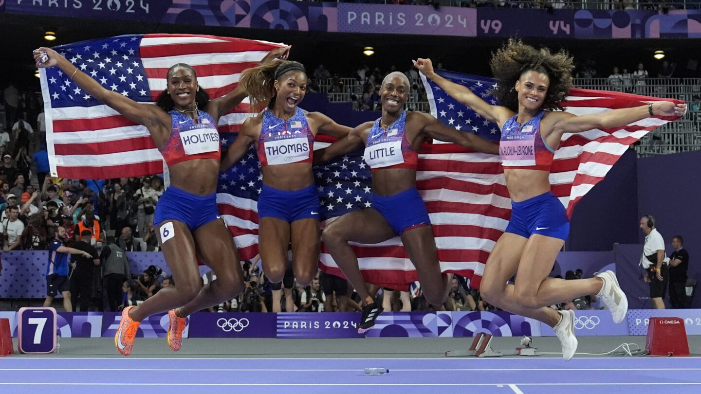 Members of the U.S. women's 4x400-meter relay team jump in celebration after winning gold, holding American flags behind them.