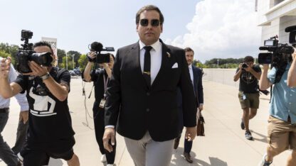 Former U.S. Rep. George Santos arrives at court in Central Islip, N.Y. He walks forward, wearing sunglasses, while cameras point at him.