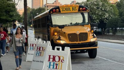 The first school bus arrives at Hope-Hill Elementary School on Aug. 1, 2024 in front of a line of signs with the school's name.