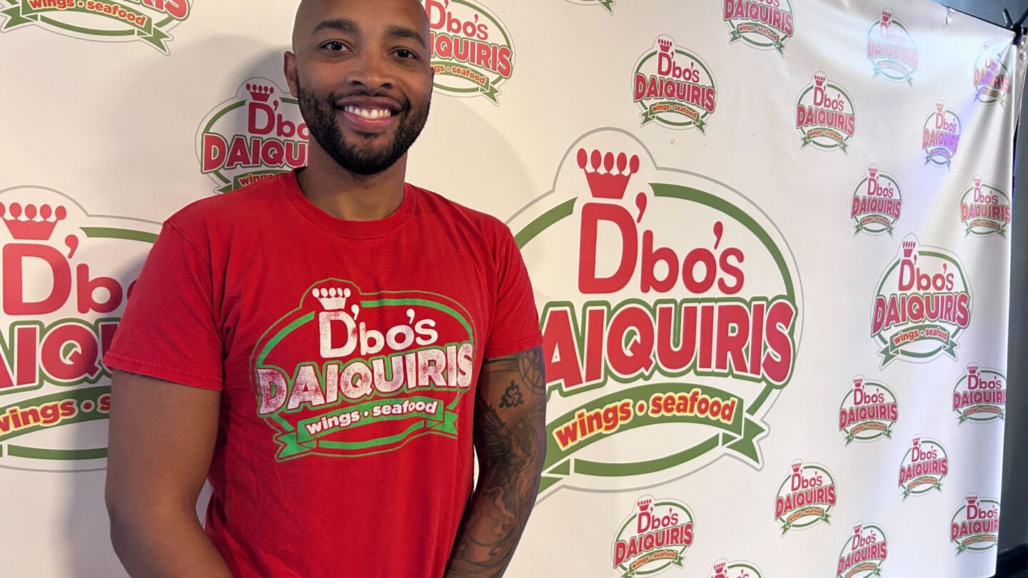 Julian Boyd wears a shirt from his restaurant, D'Bo's, while standing in front of a logo backdrop with his restaurant's name on it.