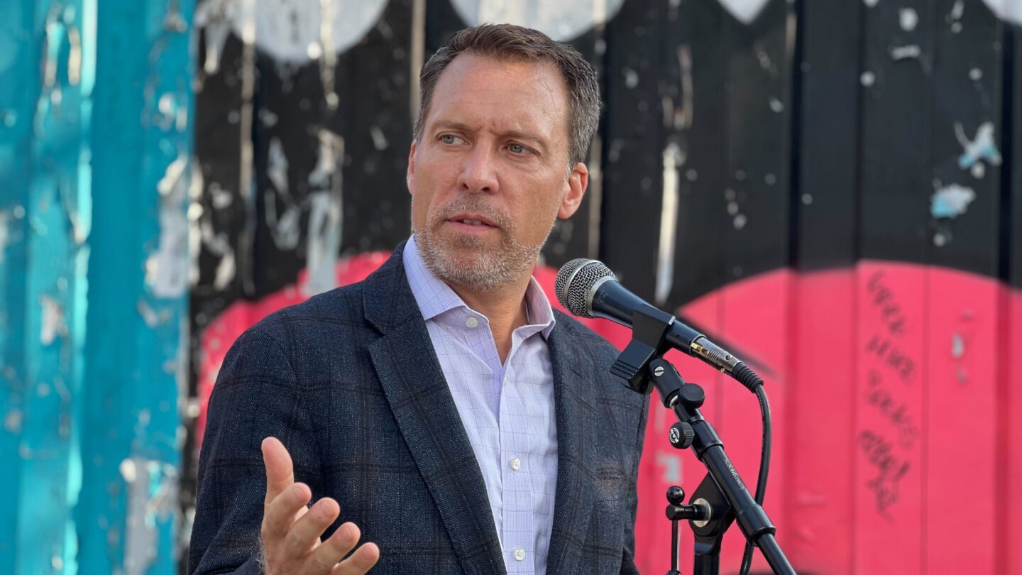 Doug Shipman raises a hand while speaking into a microphone.