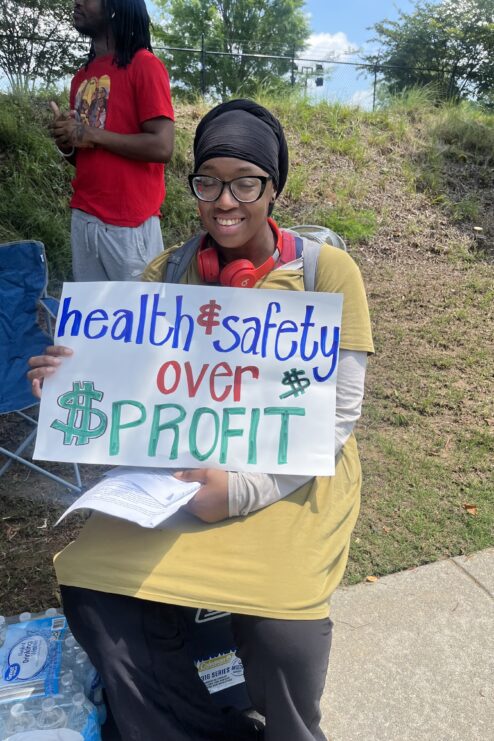 A woman in a yellow shirt holding a sign that says health and safety over profit poses for a photo.