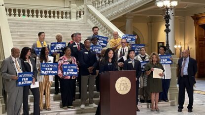 A group of people stand in front of a podium at the bottom of a staircase, holding signs that say We Demand Fair Elections