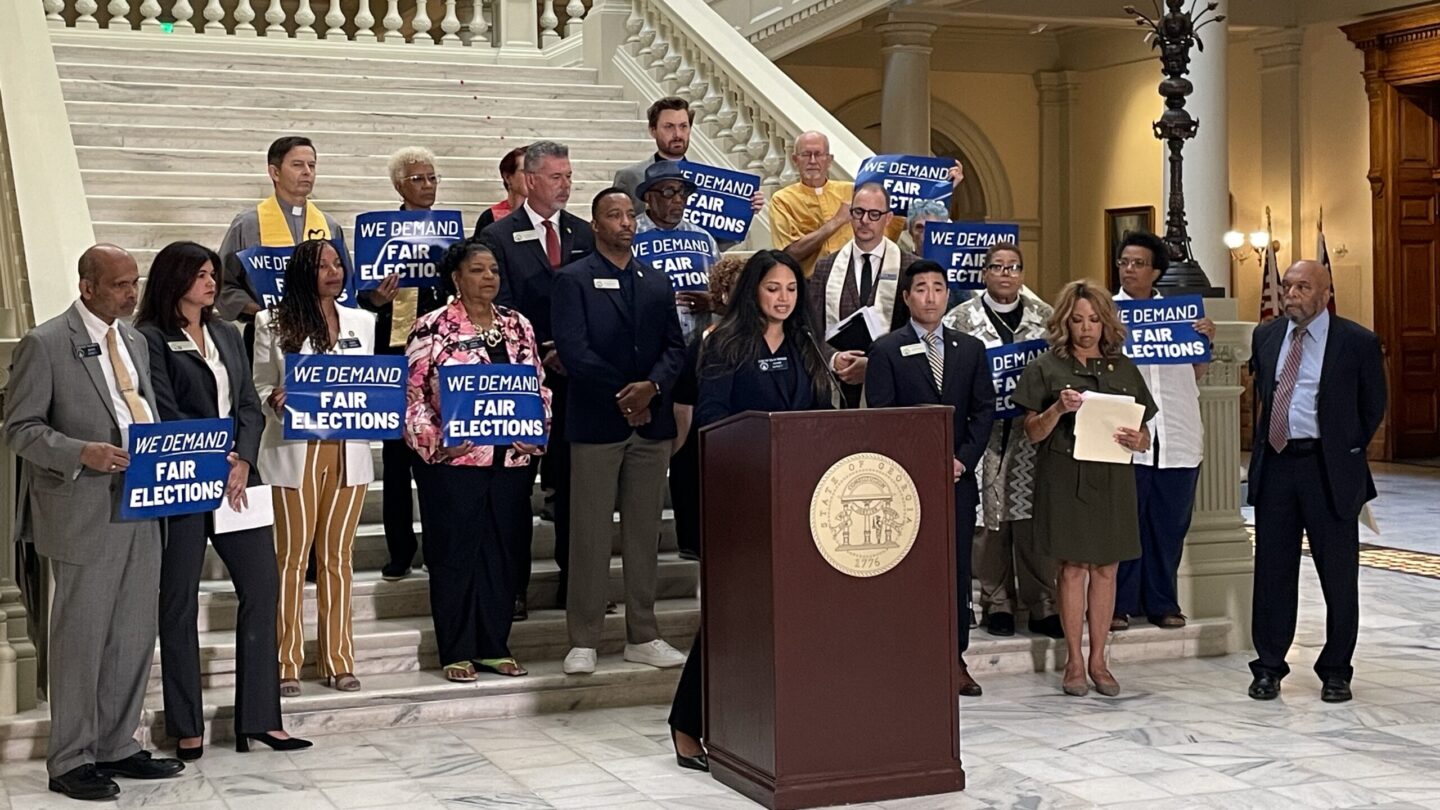 A group of people stand in front of a podium at the bottom of a staircase, holding signs that say We Demand Fair Elections