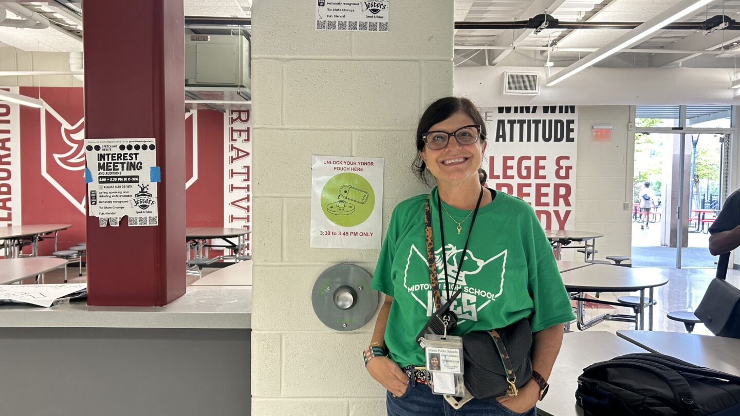 Cheryl Nahmias smiles and stands next to a magnet where students unlock their device pouches at the end of the school day.