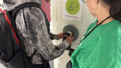 Cheryl Nahmias, wearing a green shirt, stands next to a student unlocking a device pouch.