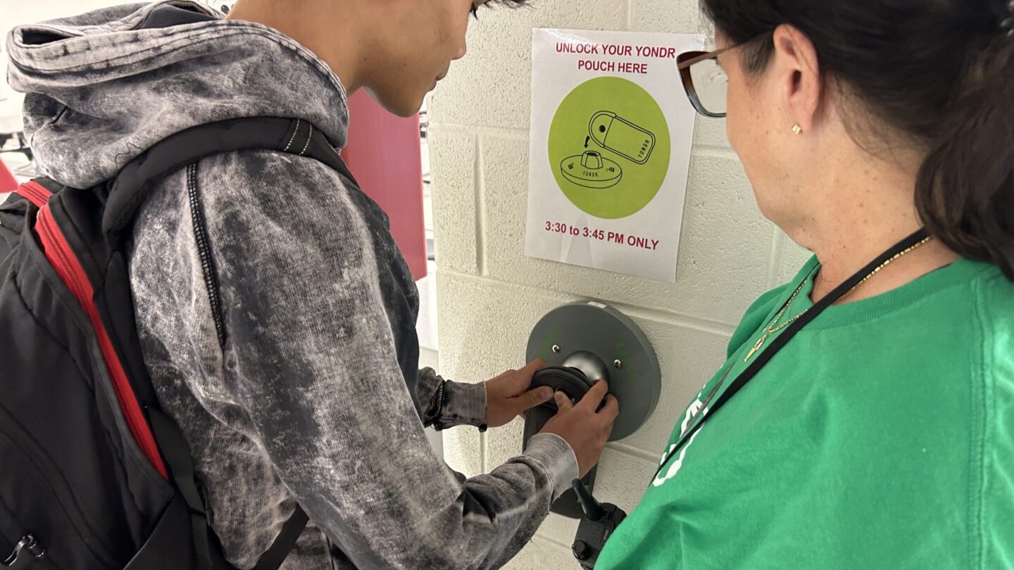 Cheryl Nahmias, wearing a green shirt, stands next to a student unlocking a device pouch.