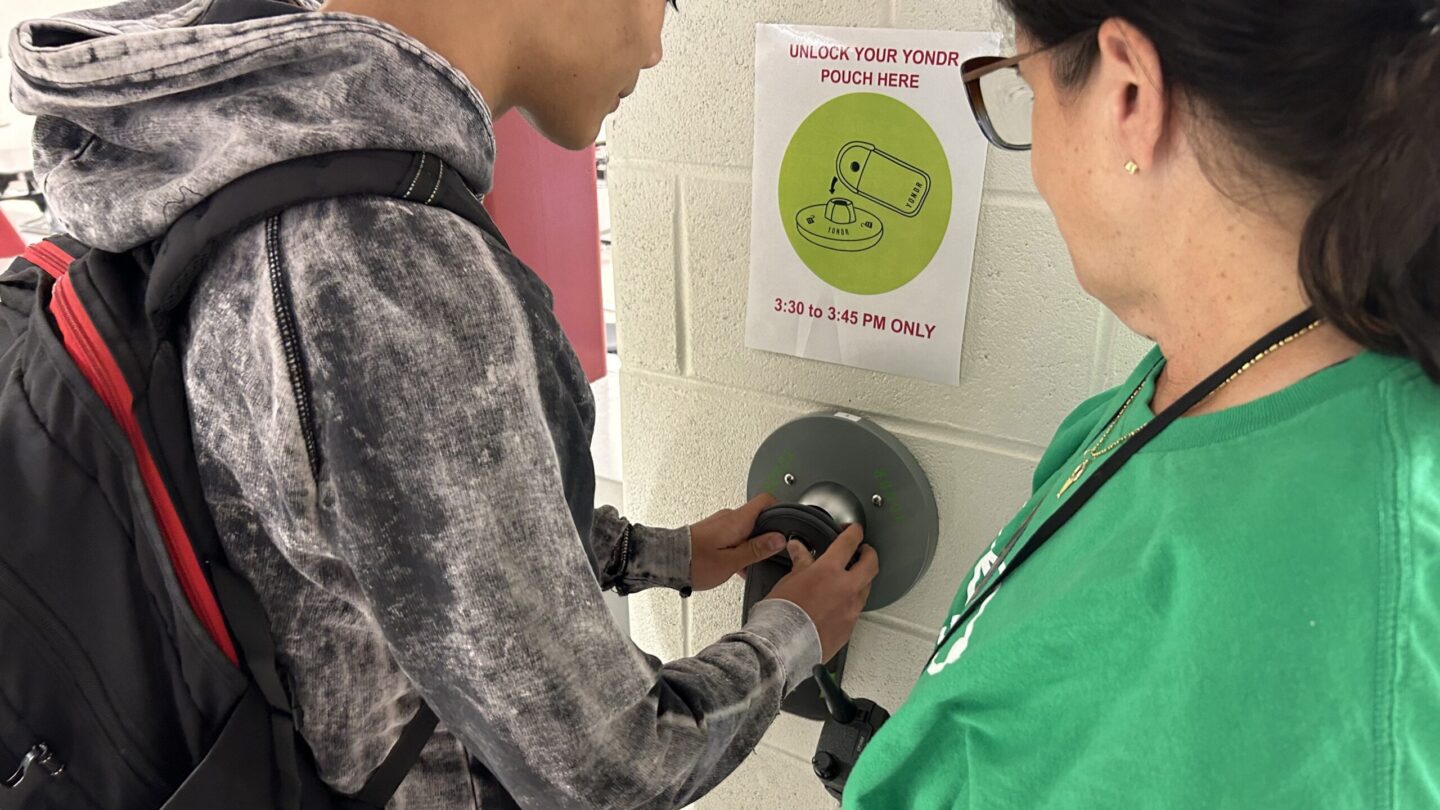 Cheryl Nahmias, wearing a green shirt, stands next to a student unlocking a device pouch.