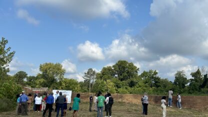 People mill around the Chattahoochee Brick Company site in front of a short, old brick wall. There's a white banner on the left about the Bipartisan Infrastructure Law.