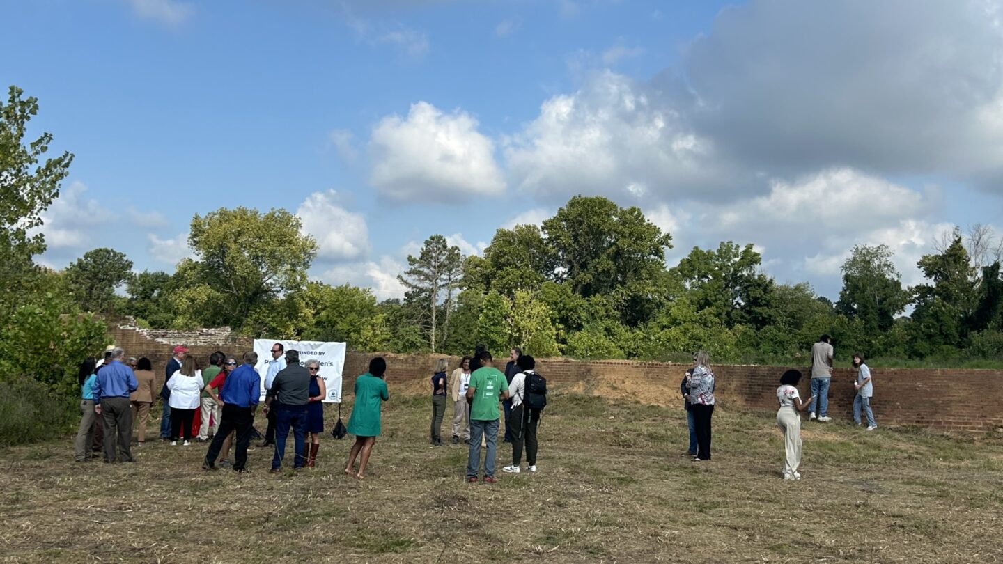 People mill around the Chattahoochee Brick Company site in front of a short, old brick wall. There's a white banner on the left about the Bipartisan Infrastructure Law.