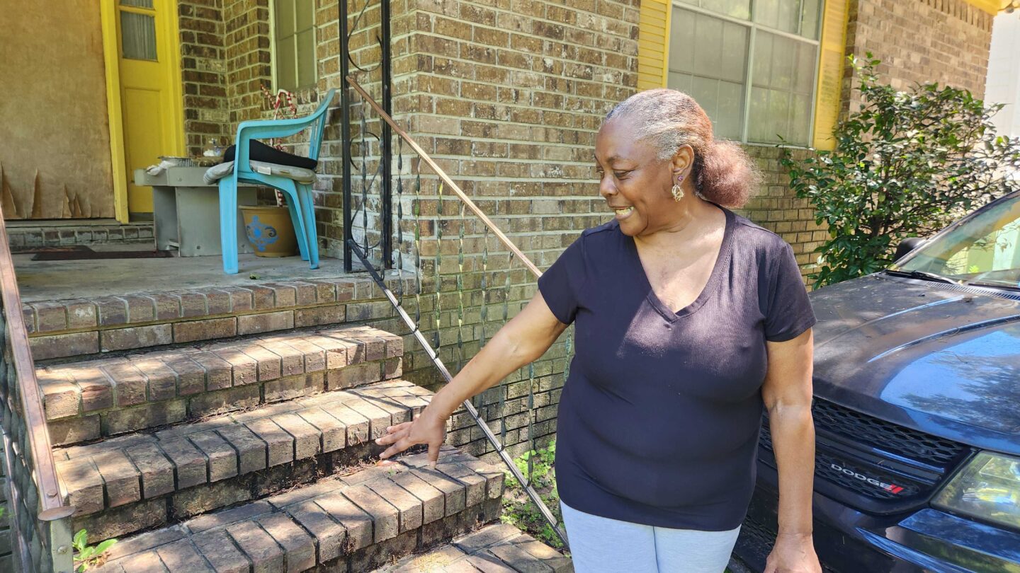 Queen Winn stands in front of her home, pointing to where flood waters from the tropical storm reached on her porch.