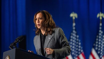 Kamala Harris stands at the podium to deliver remarks at a rally, in front of a deep blue background and several American flags.