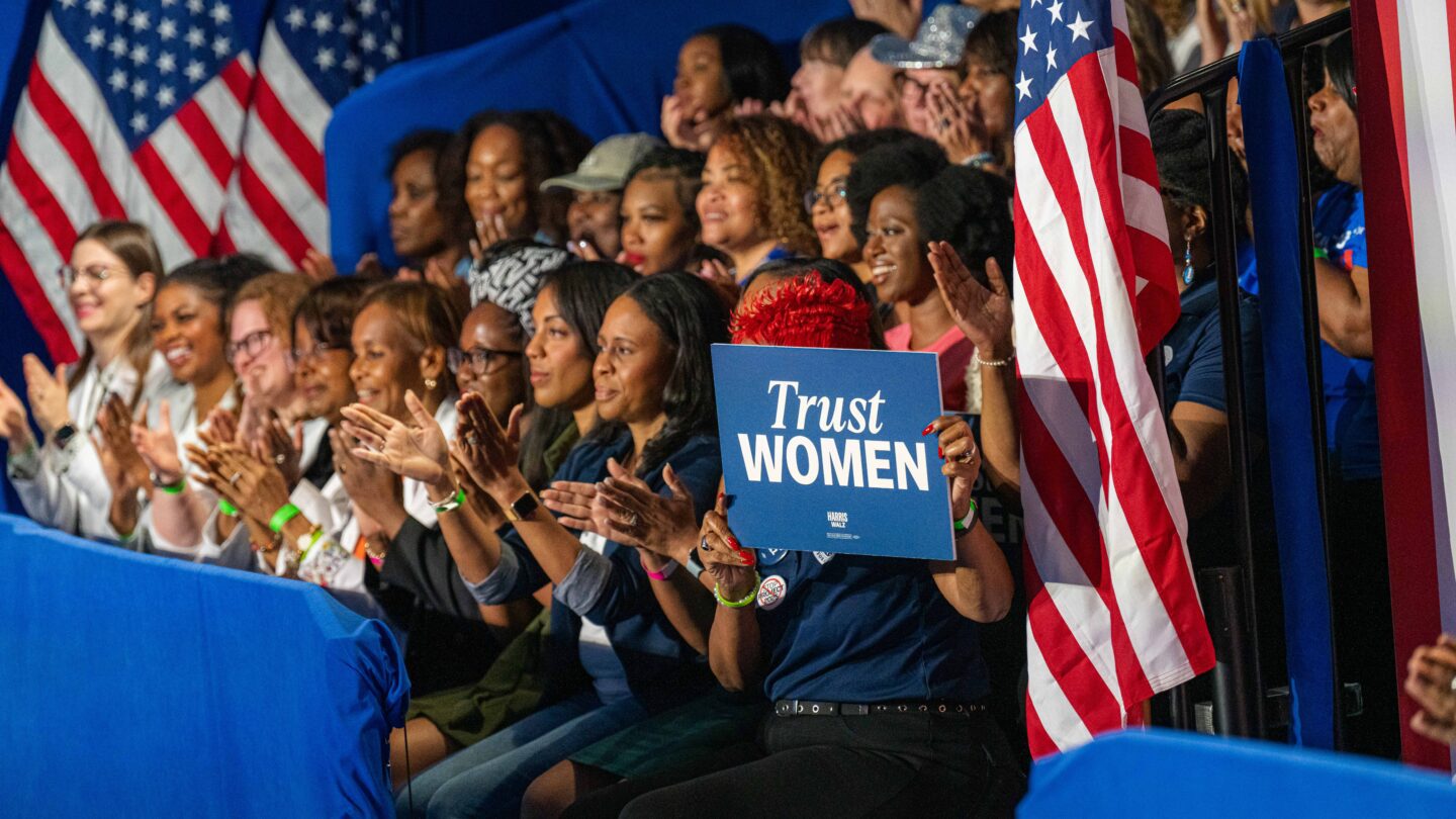 Supporters hold signs and listen and applaud at a Kamala Harris rally. The sign says Trust Women.