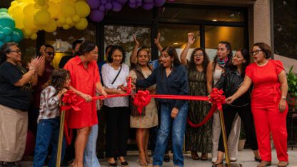 A group of people stand behind a large red ribbon as someone cuts it.