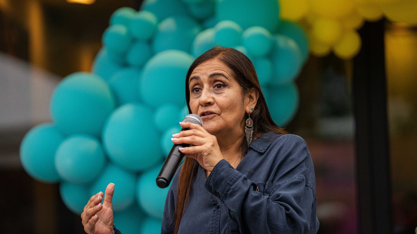 A woman stands in front of a turquoise and yellow balloon banner and speaks into a microphone.