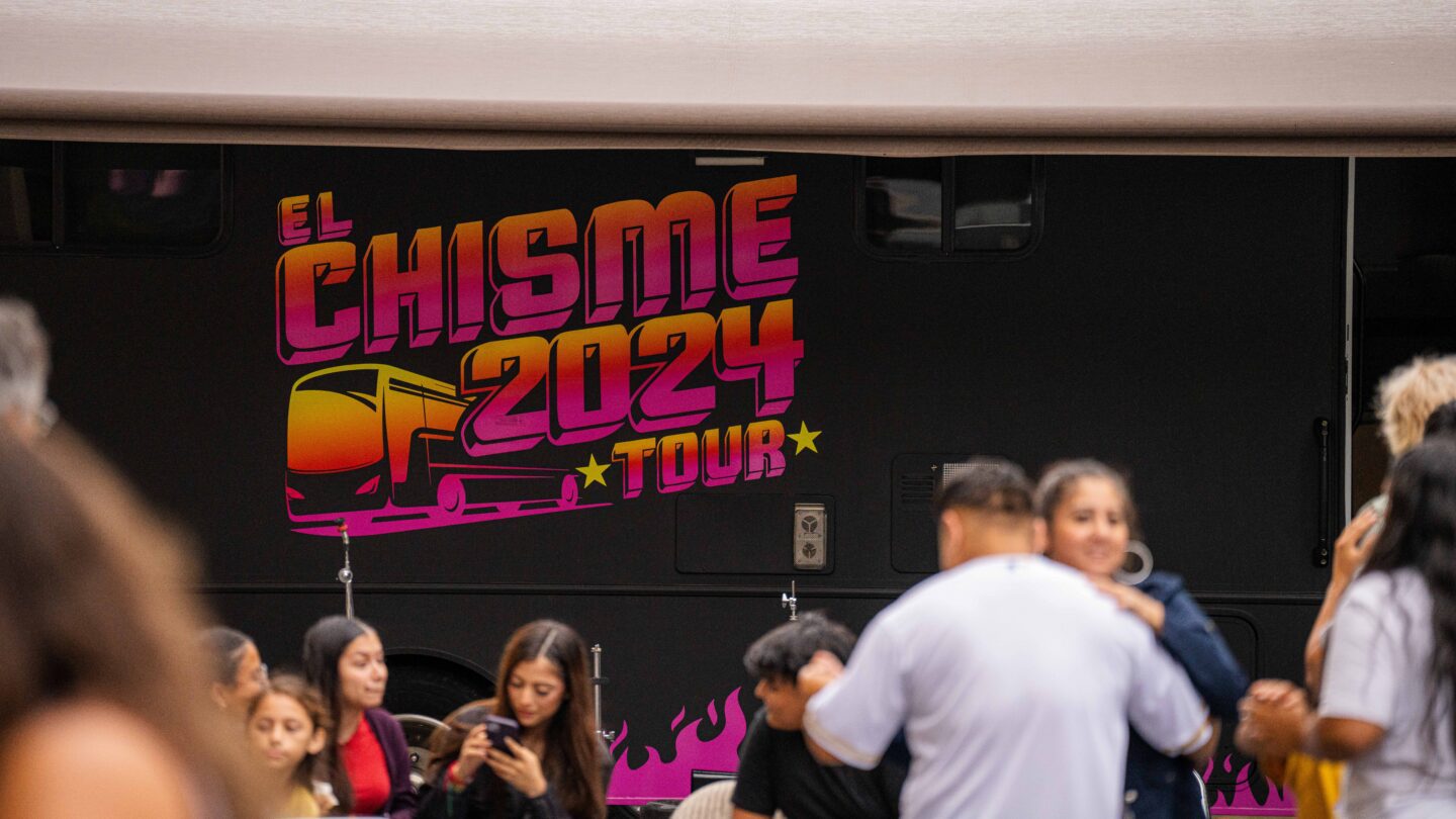 Two people dance in front of a bus with red and orange letters saying El Chisme Tour 2024