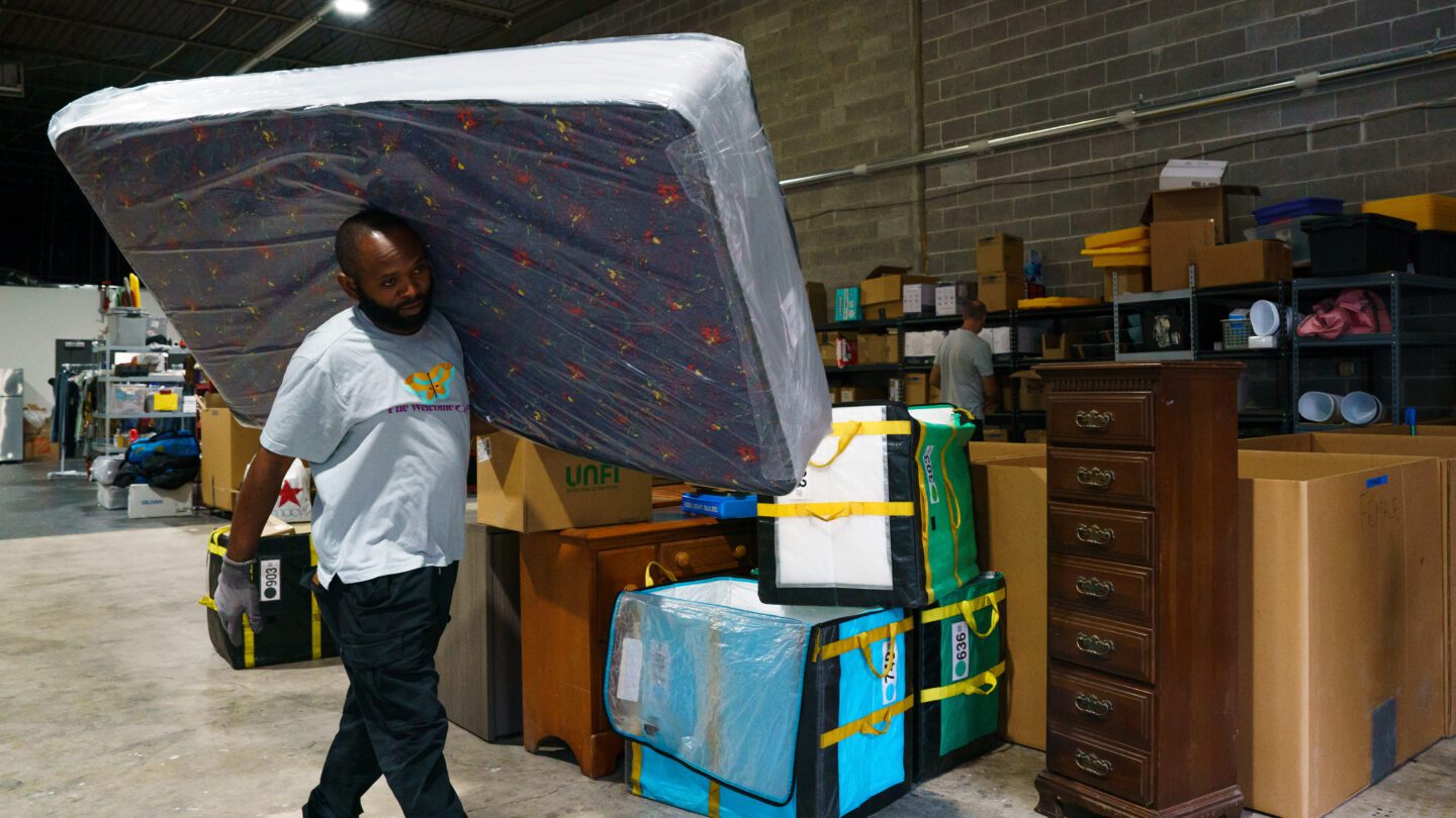 A man carries a mattress through a warehouse of other objects, including furniture.