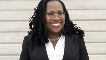 Supreme Court Justice Ketanji Brown Jackson is smiling in front of a white wall.