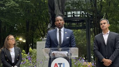 Atlanta Mayor Andre Dickens stands at a podium in front of a statue in Woodruff Park. Cathryn Vassell, left, CEO of Partners for Home, and Frank Fernandez, right, President and CEO at Community Foundation for Greater Atlanta, stand beside him.