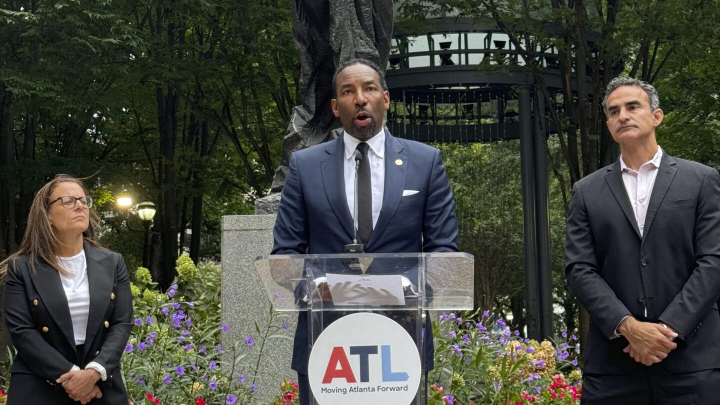 Atlanta Mayor Andre Dickens stands at a podium in front of a statue in Woodruff Park. Cathryn Vassell, left, CEO of Partners for Home, and Frank Fernandez, right, President and CEO at Community Foundation for Greater Atlanta, stand beside him.