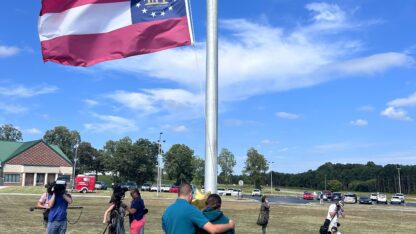 Two people stand in front of a flag pole with flowers at the bottom in mourning.