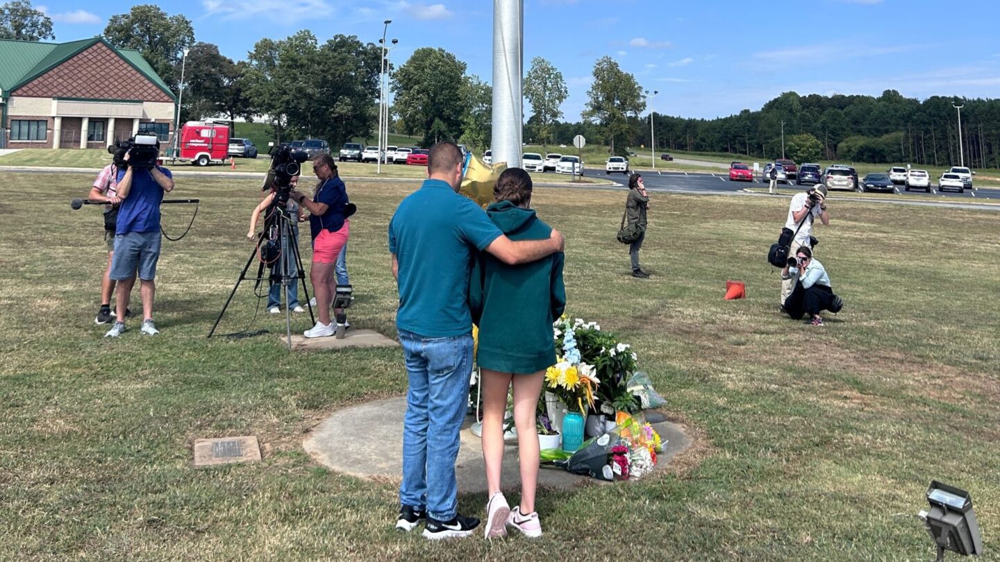 Two people stand in front of a flag pole with flowers at the bottom in mourning.