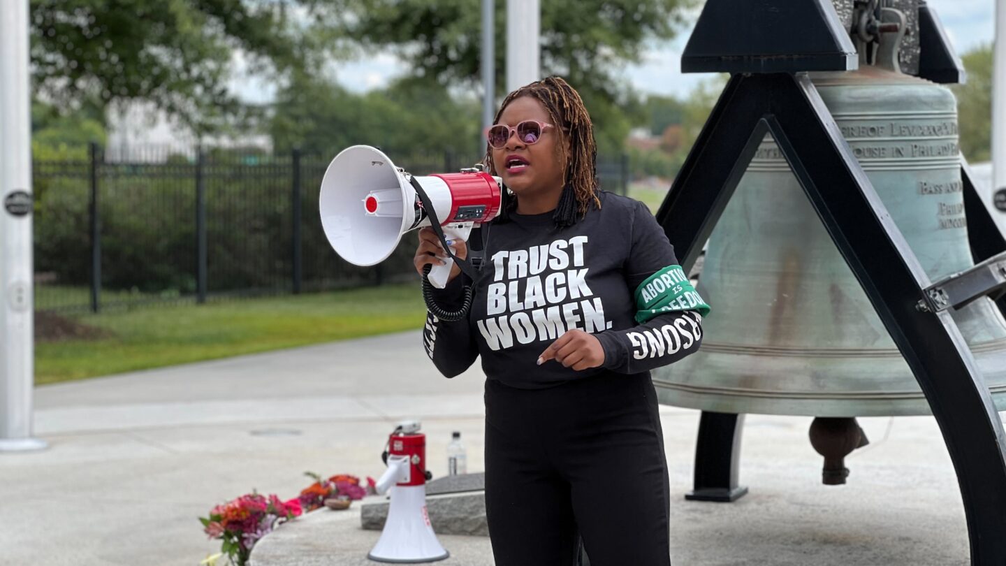 State Representative Park Cannon wearing a Trust Black Women shirt, standing in front of the Liberty Bell and holding a megaphone at a rally.